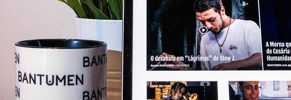 A tablet displaying a magazine website is positioned on a table, next to a mug with the word 'Bantumen' printed multiple times and a small potted plant. The tablet shows various articles and images on the screen.