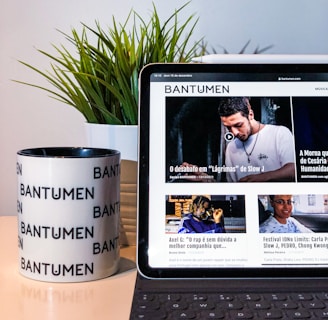 A tablet displaying a magazine website is positioned on a table, next to a mug with the word 'Bantumen' printed multiple times and a small potted plant. The tablet shows various articles and images on the screen.