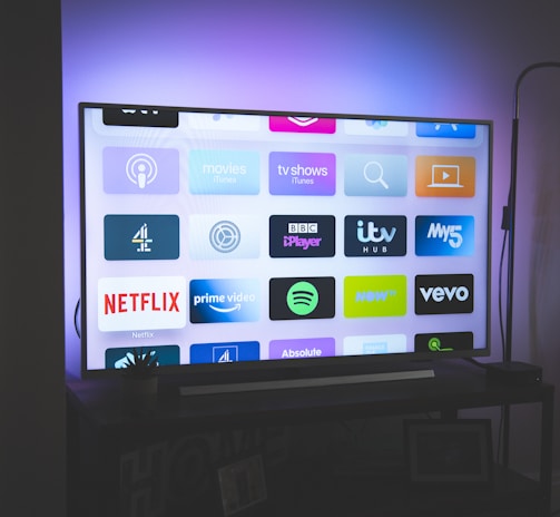 A flat-screen television is displayed in a dimly lit room, showcasing a variety of streaming service apps on its screen, such as Netflix, Prime Video, and Spotify. The TV is positioned on a black stand beside a tall floor lamp, casting a soft ambient light on the surrounding area.