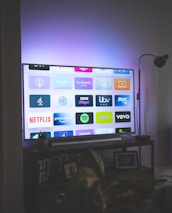 A flat-screen television is displaying a variety of streaming app icons, including Netflix, Prime Video, Spotify, and ITV Hub among others. The TV is placed on a media console with a small potted plant beside it. A tall lamp stands to the right, and a faint, ambient light casts a purple hue on the wall above the TV.