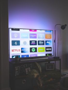 A flat-screen television is displaying a variety of streaming app icons, including Netflix, Prime Video, Spotify, and ITV Hub among others. The TV is placed on a media console with a small potted plant beside it. A tall lamp stands to the right, and a faint, ambient light casts a purple hue on the wall above the TV.