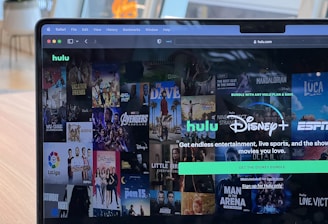 A laptop screen displays a web page featuring a mix of movie and TV show covers, highlighting popular titles and a Hulu and Disney+ bundle promotion. There are visible icons and images of films and shows alongside promotional text offering entertainment and live sports.