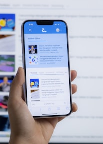 A hand holding a smartphone displaying a news website, with a blurred large computer screen in the background also showing similar content. The webpage on the phone features articles with images and text.