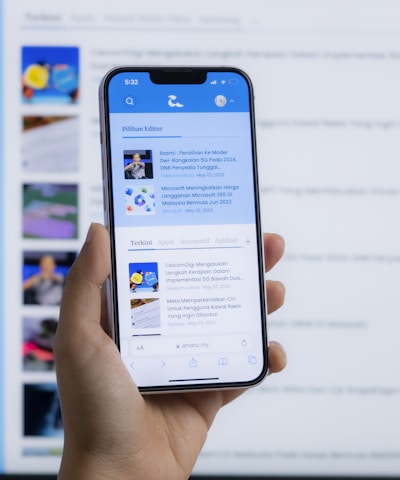 A hand holding a smartphone displaying a news website, with a blurred large computer screen in the background also showing similar content. The webpage on the phone features articles with images and text.