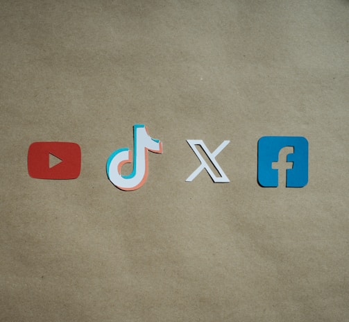 Four social media platform logos are arranged on a brown background. From left to right, the logos are for YouTube in red, TikTok in a combination of red, blue, and white, X in white, and Facebook in blue.